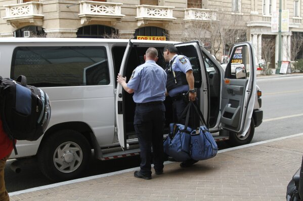 Immigration guards finish loading a van of asylum seekers leaving court under guard after some of them learned that they won't have to return to Mexico in light of a major federal court ruling against the Trump administration on Friday, Feb. 28, 2020, in El Paso, Texas.  The 9th U.S. Circuit Court of Appeals ruled Friday that sending asylum seekers to wait in Mexico is illegal nationwide. The ruling came during the asylum testimony of one family in the group being put into the van. The blue bags, marked "for court only" are typically laden with sandwiches and water bottles for migrants appearing in court. (AP Photo/Cedar Attanasio)