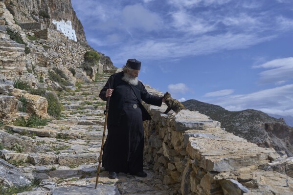 O monge cristão ortodoxo Padre Spyridon de Amorgos acaricia um gato ao sair do Mosteiro de Panagia Hozoviotissa, visto ao fundo, na Ilha de Amorgos, Grécia, no domingo, 30 de março de 2025. (Foto AP/Petros Giannakouris)