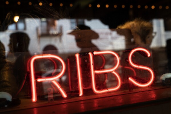 A neon sign advertises ribs at a steakhouse restaurant as pedestrians are reflected walking by Friday, April 21, 2023 in Forth Worth, Texas, Friday, April 21, 2023. The beef industry is the third-largest economic generator in Texas, contributing roughly $12 billion annually to the state's economy and employing thousands of people. (AP Photo/David Goldman)