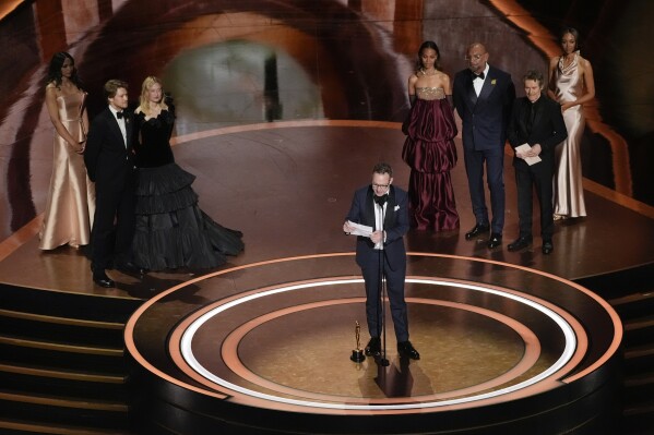 CORRECTS NAME AND AWARD - Lol Crawley accepts the award for best cinematography for "The Brutalist" during the Oscars on Sunday, March 2, 2025, at the Dolby Theatre in Los Angeles. Joe Alwyn, from left, Alba Rohrwacher, Zoe Saldana, Dave Bautista, and Willem Dafoe look on from rear of the stage. (AP Photo/Chris Pizzello)