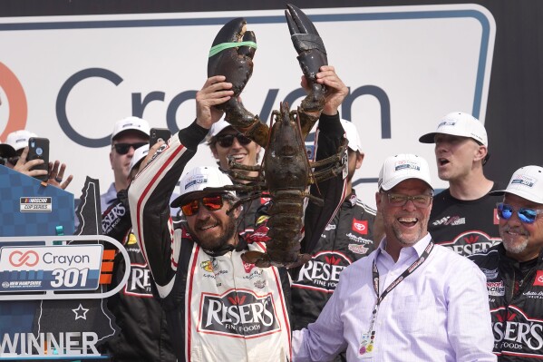 Martin Truex Jr., front left, holds up a lobster while celebrating after his win in the Crayon 301 NASCAR Cup Series race as David McGrath, second from front right, executive vice president and general manager of New Hampshire Motor Speedway, smiles Monday, July 17, 2023, at the speedway in Loudon, N.H. (AP Photo/Steven Senne)