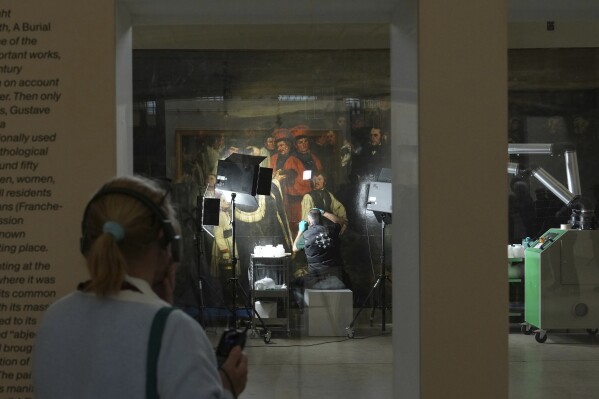 A woman watches art restorers working at the painting "A burial at Ornans" by Gustave Courbet, from behind a glass window at the Orsay museum, Wednesday, May 7, 2025 in Paris. (AP Photo/Aurelien Morissard)