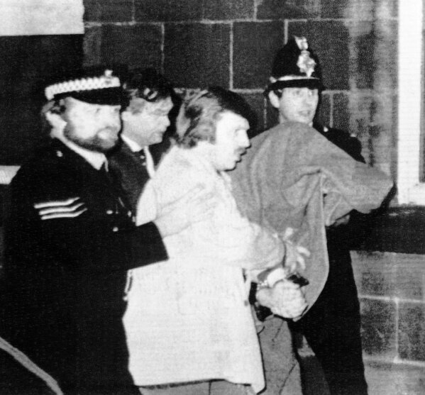 FILE - This Jan. 5, 1981 file photos shows Peter William Sutcliffe, 35, under a blanket at right, being led from Dewsbury Magistrates Court in Dewsbury by police officers. On Friday, Nov. 13, 2020, Britain’s Prison Service said that serial killer Peter Sutcliffe _ known as the "Yorkshire Ripper" died in the hospital. Sutcliffe was serving a life sentence after being convicted of murdering 13 women in northern England between 1975 and 1980. (AP Photo/Pyne, file)