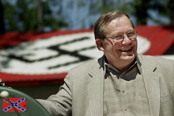 
              FILE - In this May 22, 2001, file photo, Norm Gissel smiles as he talks about the imminent dismantling of the former headquarters of the Aryan Nations in Hayden Lake, Idaho. Behind Gisel is a Nazi insignia painted atop the roof of the compound's cafeteria. Nearly two decades after the Aryan Nations compound was demolished in Idaho, far-right extremists are maintaining a presence in the Pacific Northwest. (AP Photo/Elaine Thompson, File)
            