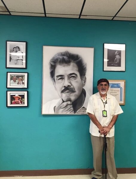In this April 2016, photo provided by Irene Monica Sanchez, civil rights leader Bobby Lee Verdugo stands in front of a portrait of Chicano Movement leader Rodolfo "Corky" Gonzales at Escuela Tlatelolco in Denver, Colo. Verdugo, one of the leaders of the 1968 East Los Angeles high school walkout to protest discrimination and dropout rates among Mexican American students, died at age 69, on Friday, May 1, 2020. His daughter, Monica Verdugo, announced on Facebook that her father died peacefully surrounded by his wife, Yoli Rios, and his family. (Courtesy Irene Monica Sanchez via AP)