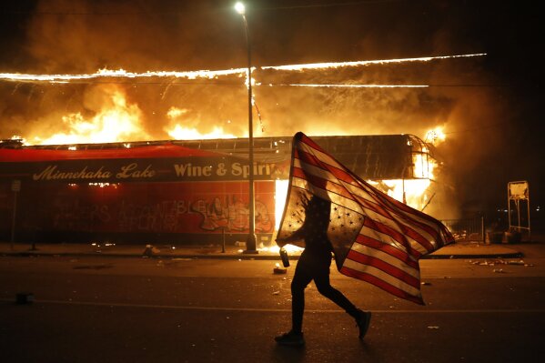 FILE - In this May 28, 2020, file photo, a protester carries a American flag upside down, a sign of distress, next to a burning building in Minneapolis. Protests over the death of George Floyd, a black man who died in police custody, broke out in Minneapolis for a third straight night. (AP Photo/Julio Cortez, File)