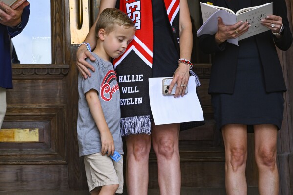 Noah Neumann, 6, is comforted by his Covenant mother as families engage in a prayer service leading up to a special session of the state legislature before a news conference at the state capitol, Thursday, July 20, 2023, in Nashville, Tenn. The Covenant School in Nashville was subject to a mass shooting which killed three students and three staff on March 27. The first names of children killed in the shooting are displayed on the wardrobe of the mother. (AP Photo/John Amis)