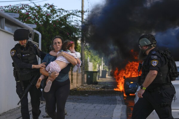 Police officers evacuate a woman and a child from a site hit by a rocket fired from the Gaza Strip, in Ashkelon, southern Israel, Saturday, Oct. 7, 2023. The rockets were fired as Hamas announced a new operation against Israel. (AP Photo/Tsafrir Abayov)