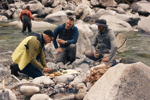 This image released by National Geographic shows Stanley Tucci, left, preparing a dish for anglers on the banks of the Sarca River during a visit to Italy's northernmost region, during the filming of his series "Tucci in Italy." (Matt Holyoak/National Geographic via AP)