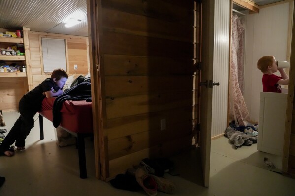 Hunter Prince, left, watches a video on his computer inside his mother and stepfather's bedroom as his little brother, Waylon, right, finishes brushing his teeth at Camp Graves, a nonprofit helping provide temporary housing, Tuesday, Jan. 16, 2024, in Water Valley, Ky. Two years after the tornado outbreak that killed dozens and leveled much of the real estate in Mayfield, many people, like the Prince family, are still living through another, slower disaster, the search for housing. (AP Photo/Joshua A. Bickel)