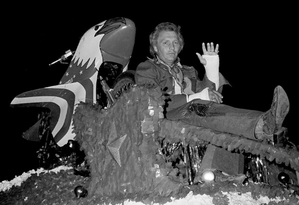With his injured leg propped up and both arms in a cast, American daredevil stunt motorcyclist and Mardi Gras Grand Marshal, Evel Knievel, manages a small wave as he rides in a carnival procession in New Orleans, Feb. 13, 1977. Knievel, who was injured recently in Chicago during a practice jump, was unable to throw trinkets and doubloons from his float because of his injuries. (AP Photo, file)