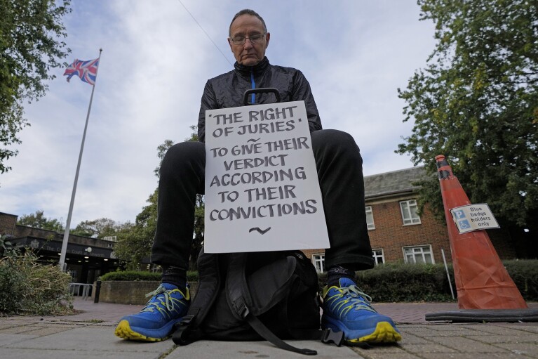 A protestor shows a placard as he sits outside the entrance to Isleworth Crown Court in London, Friday, Oct. 6, 2023. (AP Photo/Frank Augstein)