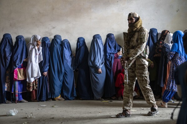 FILE - A Taliban fighter stands guard as women wait to receive food rations distributed by a humanitarian aid group, in Kabul, Afghanistan, on May 23, 2023. A U.N. report says the Taliban are restricting Afghan women's access to work, travel and healthcare if they are unmarried or don't have a male guardian, a mahram. The Taliban have banned women from education, most jobs and public spaces like parks since seizing control of Afghanistan in 2021. (AP Photo/Ebrahim Noroozi, File)