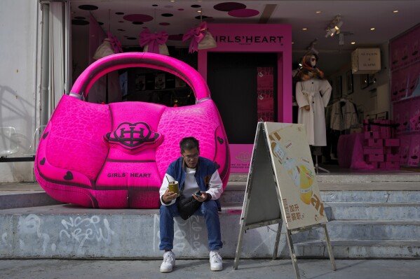 A man browses his smartphone as he rests near a giant handbag on display outside a fashion boutique in Xiamen in southeast China's Fujian province on Dec. 30, 2023. A survey of factory managers in China shows manufacturing contracted in December in the latest sign the world's second-largest economy remains sluggish. (AP Photo/Andy Wong)