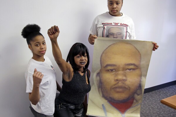 FILE - Hope Coleman raises her fist as she poses with her granddaughters, Danina, left, and Destiny, holding a photo of her son, Terrence, April 4, 2018, in Boston. Coleman filed a federal lawsuit in 2018 accusing the city of failing to properly train officers on how to deal with mentally ill people. Terrence Coleman, who was mentally ill, was killed by police in 2016 after his mother called 911 for an ambulance to take him to the hospital. The city of Boston will pay about $4.6 million to settle a wrongful death lawsuit. (AP Photo/Elise Amendola, File)