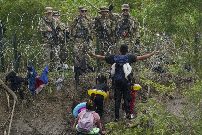 A migrant standing behind razor wire on the banks of the Rio Grande River, seen from Matamoros, Mexico, on May 11, 2023, gestures to Texas National Guardsmen.  (AP Photo/Fernando Llano, File)