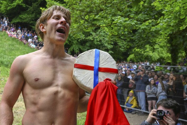 Competitor Tom Kopke of Munich, Germany, holds the cheese after wiining the men's downhill race category of the traditional annual Cheese Rolling contest at Cooper's Hill in Brockworth, Gloucestershire, England, Monday, May 26, 2025.(AP Photo/Anthony Upton)