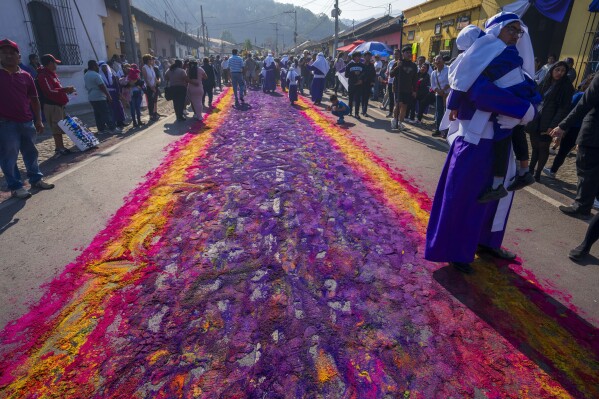 A penitent knows as a cucurucho holds a child standing on the edge of a sawdust carpet constructed by the Alvarez family and friends, after a Holy Week procession walked over it in Antigua, Guatemala, Friday, March 29, 2024. (AP Photo/Moises Castillo