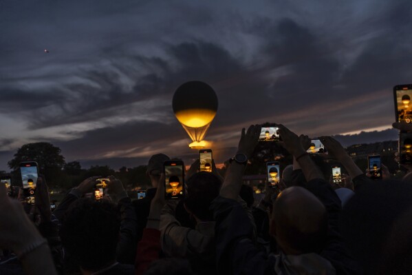 A crowd takes photos at the gates to the Tuileries Garden as the cauldron rises at sunset during the 2024 Summer Olympics, Saturday, July 27, 2024, in Paris. (AP Photo/David Goldman)