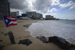 FILE - In this May 21, 2020 file photo, a Puerto Rican flag flies on an empty beach at Ocean Park, in San Juan, Puerto Rico. Puerto Rico’s governor announced Thursday, June 11, that she will lift nearly all restrictions aimed at curbing coronavirus cases, which means beaches, churches and businesses including movie theaters and gyms across the U.S. territory will reopen after three months. (AP Photo/Carlos Giusti, File) PUERTO RICO OUT-NO PUBLICAR EN PUERTO RICO