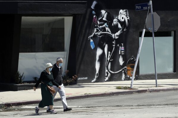 People wearing masks walk past a mural depicting a coronavirus theme during the COVID-19 pandemic Tuesday, April 14, 2020, in Los Angeles. California Gov. Gavin Newsom on Tuesday unveiled an outline for what it will take to lift coronavirus restrictions in the nation's most populous state, asking more questions than answering them as he seeks to temper the expectations of a restless, isolating public. (AP Photo/Marcio Jose Sanchez)