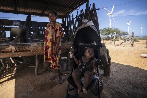 An Indigenous woman from the Wayuu community cooks near her baby as wind turbines operate in the distance on the outskirts of Cabo de la Vela, Colombia, Feb. 7, 2025. (AP Photo/Ivan Valencia, File)