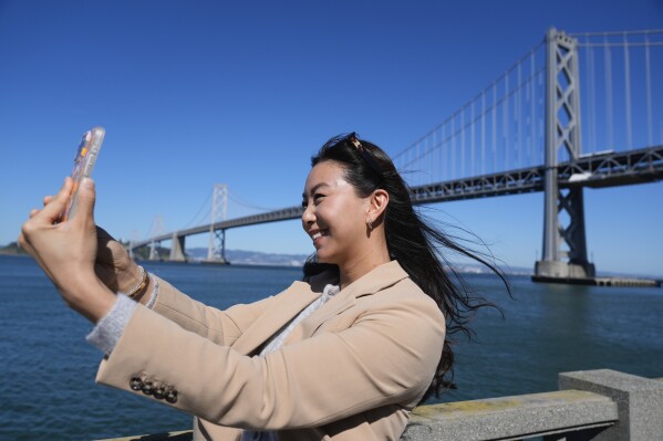 Content creator Cynthia Huang Wang works below the San Francisco-Oakland Bay Bridge in San Francisco, Monday, April 8, 2024. Despite a strong job market, there are still thousands of people who have found themselves out of work across industries stretching from tech to retail to media. But rather than trying to find another job in their old role, some workers are turning to online content creation. (AP Photo/Eric Risberg)