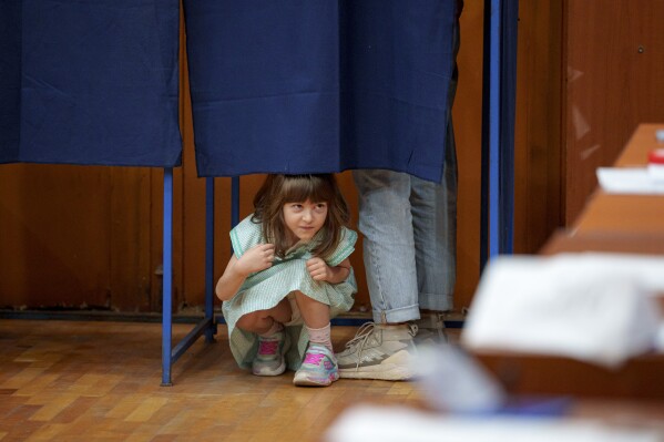 A little girl peers from under a curtain as her father casts his vote in a voting booth in the first round of the presidential election redo in Bucharest, Romania, Sunday, May 4, 2025. (AP Photo/Andreea Alexandru)