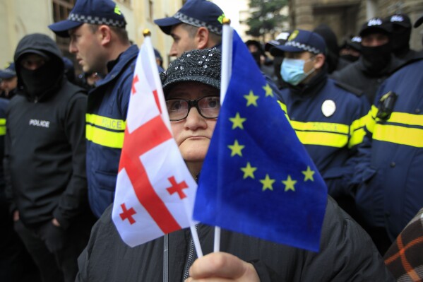 A woman with a EU and a Georgian national flags stands in front of police blocking a street during a rally to demand new parliamentary elections in the country, near the Parliament's building in Tbilisi, Georgia, on Monday, Nov. 25, 2024. (AP Photo/Shakh Aivazov)