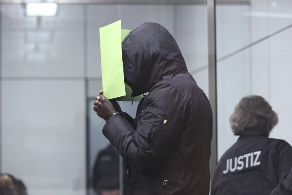 The Gambian defendant, identified only as Bai L. in line with German privacy rules, holds a folder in front of his face at the Celle Higher Regional Cour in Celle, Germany, Monday, April 25, 2022. The man went on trial in Germany on Monday for his alleged role in the killing of government critics in the West African country over 15 years ago. (Ronny Hartmann/Pool Photo via AP)