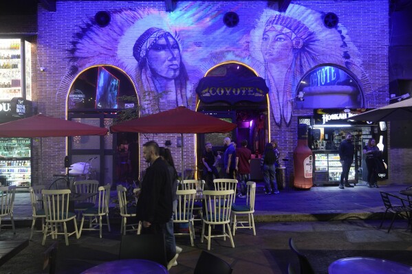 A couple strolls past the Coyote nightclub in Lleras Park in Medellin, Colombia, Friday, Feb. 2, 2024. A tourism boom in Medellin has presented officials with a new set of challenges, including an uptick in sex trafficking and the killing of tourists and Colombian women after rendezvous on dating apps. (AP Photo/Fernando Vergara)