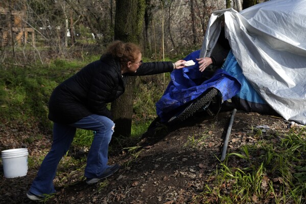 Resiliency Empowerment Support Team (REST) team member Beth Perkins, give a homeless person a hand warmer during a visit to a homeless camp in Chico, Calif., Feb. 8, 2024. A measure aimed at transforming how California spends money on mental health will go before voters in March as the state continues to grapple an unabated homelessness crisis. The REST Program does daily visits to homeless encampments to get them into treatment or housing. Butte County officials fear the REST program would lose its funding if California voters approve Proposition 1.(AP Photo/Rich Pedroncelli)