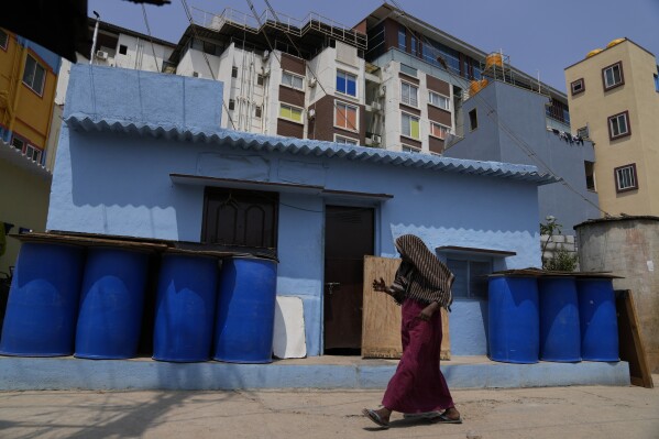 A resident of Ambedkar Nagar, a low-income settlement in the shadows global software companies in Whitefield neighborhood, walks past a row of empty barrels used to store water, in Bengaluru, India, Monday, March 11, 2024. (AP Photo/Aijaz Rahi)