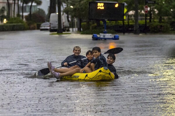 Matthew Koziol, Matías Ricci, Manuel Ricci and Raúl Fernández travel by raft through a flooded street caused by heavy rain on North Bay Road in Sunny Isles Beach, Fla., Wednesday, June 12, 2024. (David Santiago/Miami Herald via AP)