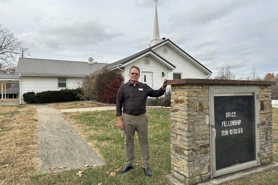 Pastor Kenny Batson stands near a sign displaying the worship service times of Grace Fellowship Church on Nov. 16, 2023, in El Dorado Springs, Mo. Batson was convicted of a series of crimes in the 1990s but became a Christian pastor after being released from prison. He was pardoned by Missouri Gov. Mike Parson. (AP Photo/David A.Lieb)