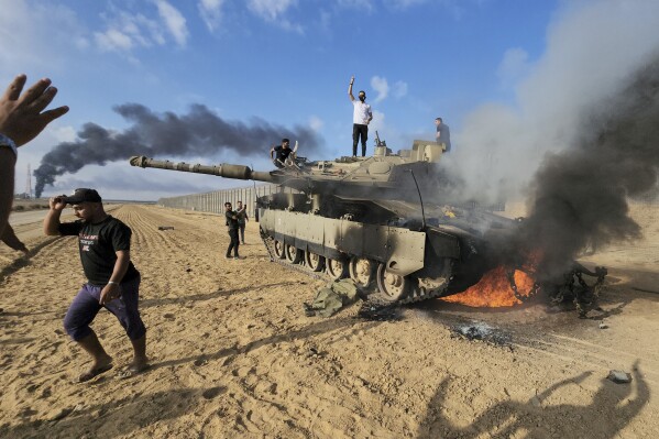 Palestinians celebrate by a destroyed Israeli tank at the border fence between Israel and the Gaza Strip, east of Khan Younis, during a surprise attack on Israel, Saturday, Oct. 7, 2023. (AP Photo/Hassan Eslaiah)