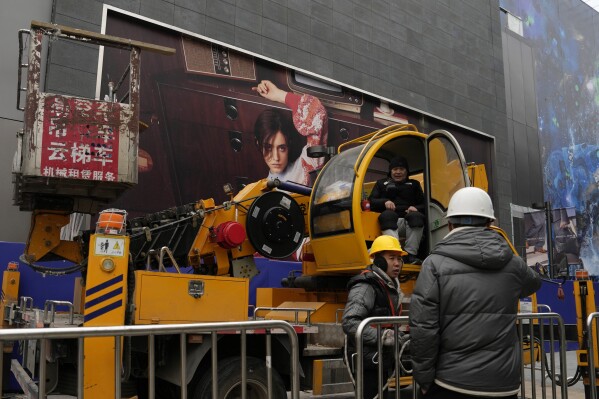 Men work with a crane near an advertisement at a shopping mall in Beijing, Wednesday, Jan. 17, 2024. China's economy for the October-December quarter grew at a quicker rate, allowing the Chinese government to hit its target of about 5% annual growth for 2023 even though trade data and the economic recovery remain uneven. (AP Photo/Ng Han Guan)