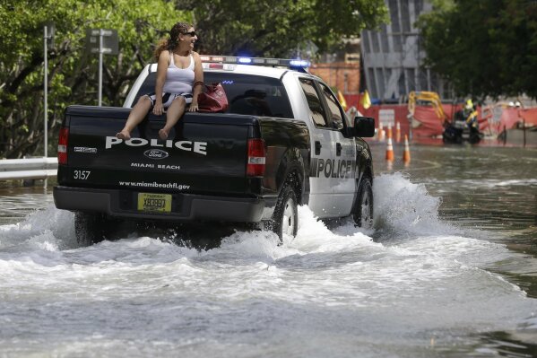 FILE- In this Sept. 30, 2015 file photo, a woman gets a ride on a police truck navigating a flooded street in Miami Beach, Fla. The street flooding was in part caused by high tides due to the lunar cycle, according to the National Weather Service. When Democratic presidential candidates meet in Miami for their first debate it'll be in what you could call the country's Ground Zero for any climate-related sea level rise. (AP Photo/Lynne Sladky, File)