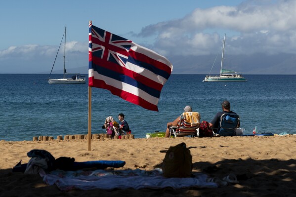 People sit on Kaanapali Beach in front of a flag of Hawaii planted in the sand, Wednesday, Dec. 6, 2023, in Lahaina, Hawaii. Residents and survivors still dealing with the aftermath of the August wildfires in Lahaina have mixed feelings as tourists begin to return to the west side of Maui. (AP Photo/Lindsey Wasson)
