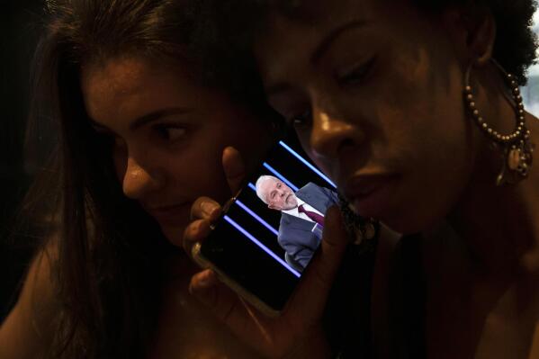 FILE - Women listen on a mobile phone to an interview of former president Luiz Inacio Lula da Silva, who is running again for president, with Jornal Nacional on TV Globo, outside a bar in Rio de Janeiro, Brazil, Thursday, August 25, 2022. The Superior Electoral Court, the country's top electoral authority, announced Thursday, Oct, 20, that it would be banning "false or seriously decontextualized" content that “affects the integrity of the electoral process.” No request from a prosecutor or complainant is necessary for the court to take action. (AP Photo/Bruna Prado, File)