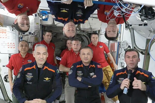 In this image from video provided by NASA, the 11 International Space Station crew members representing Expedition 70 (red shirts) and Axiom Space 3 (dark blue suits) crews gather for a farewell ceremony calling down to mission controllers on Earth on Friday, Feb. 2, 2024. Front row from left are Italy's Walter Villadei, Turkey's Alper Gezeravci and Sweden's Marcus Wandt. Above them hanging upside down in blue is Axiom Space's Michael Lopez-Alegria, a former NASA astronaut. (NASA via AP)