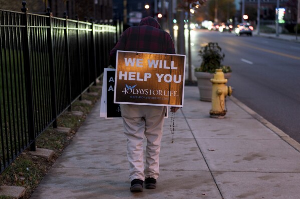 Mike Logsdon's sign catches a street light as he prays a rosary and walks in front of Planned Parenthood in Cincinnati, early Wednesday, Nov. 1, 2023. Logsdon sign reads "We Will Help You" from the 40 Days for Life campaign to end abortion. (AP Photo/Carolyn Kaster)