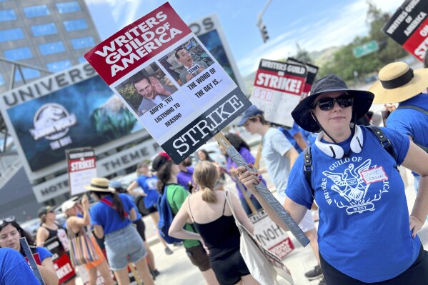 Writer Julie Benson holds a picket sign with an image of the late actor and comic Paul Reubens, dressed as his character Pee-wee Herman, outside Universal Studios on Tuesday, Aug. 1, 2023, in Universal City, Calif. The actors strike comes more than two months after screenwriters began striking in their bid to get better pay and working conditions. (AP Photo/Rick Taber)