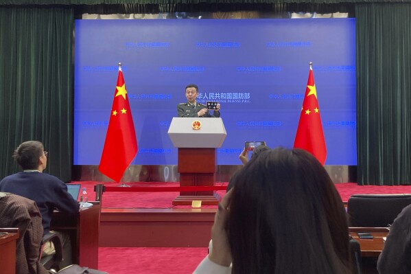 Senior Col. Wu Qian, the defense ministry's top spokesperson, holds up souvenir pins to mark the Chinese Air Force anniversary during a press conference in Beijing, Thursday, Nov. 30, 2023. China's defense ministry said Thursday that the U.S. should stop interfering in both Taiwan and the South China Sea, calling U.S. arms sales to Taiwan a powder keg that is making the situation more dangerous. (AP Photo/Ken Moritsugu)