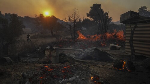 A man on a motorcycle looks on as a shipyard which is on fire in Mandra west of Athens, on Tuesday, July 18, 2023. In Greece, where a second heatwave is expected to hit Thursday, three large wildfires burned outside Athens for a second day. Thousands of people evacuated from coastal areas south of the capital returned to their homes Tuesday when a fire finally receded after they spent the night on beaches, hotels and public facilities. (AP Photo/Petros Giannakouris)