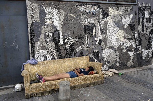 A woman sleeps on sofa that was discarded on the sidewalk next to a mural depicting Pablo Picasso's "Guernica" painting in the Padre Carlos Mugica neighborhood of Buenos Aires, Argentina, Thursday, Dec. 14, 2023. (AP Photo/Rodrigo Abd)