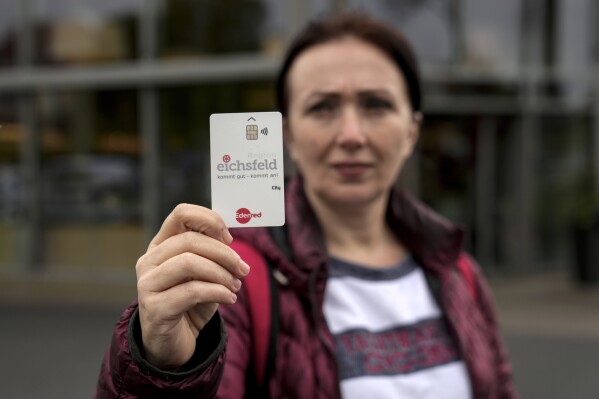 Erdina Laca, a 45-year-old asylum seeker, shows her special payment card in front of a grocery store, in Eichsfeld, Germany, Wednesday, April 24, 2024. Across Germany, cities and counties are introducing new payment cards for asylum-seekers. The new rule, which was passed by parliament last month, calls for the migrants to receive their benefits on a card that can be used for payments in local shops and services. (AP Photo/Ebrahim Noroozi)