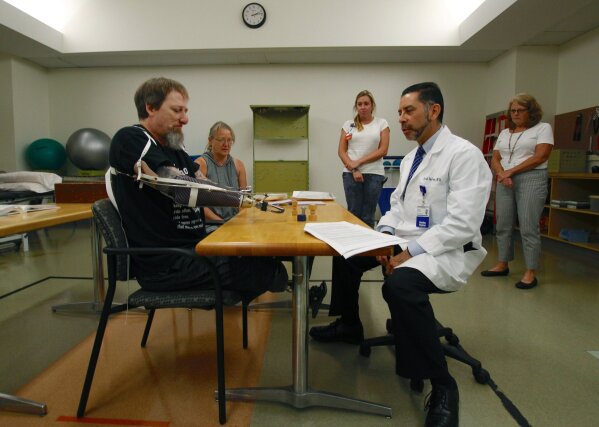 In this Aug. 19, 2019 photo, Greg Manteufel tries out a new prosthetic arm during occupational therapy at Froedtert & the Medical College of Wisconsin in Milwaukee, as he talks to Dr. David Del Toro, the medical director for inpatient rehab. Manteufel lost parts of his arms and legs, as well as the skin of his nose and part of his upper lip from capnocytophaga, a bacteria commonly found in the saliva or cats and dogs that almost never leads to people getting sick, unless the person has a compromised immune system. He was perfectly healthy when he got sick in June of 2018. Over the last seven years, a team of researchers at Brigham and Women's Hospital in Boston, connected to Harvard Medical School, have tested other healthy people who were affected and developed a theory on why they were affected- a gene change in all the victims. (AP Photo/Carrie Antlfinger)