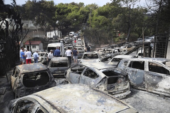 People stand amid the charred remains of burned-out cars in Mati east of Athens, Tuesday, July 24, 2018. (AP Photo/Thanassis Stavrakis, File)