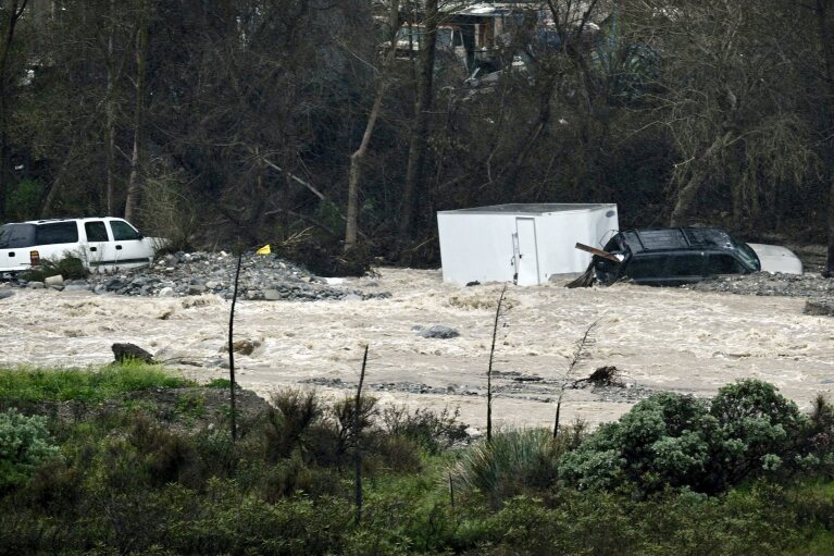 Two vehicles, one towing a trailer, sit in the rocks on the bank of a swollen Cajon Creek near Devore, Monday, Feb. 5, 2024, after being swept away in the floodwaters the night before. (Watchara Phomicinda/The Orange County Register via AP)